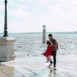 A father joyfully swings his daughter by the sea on a bright day, capturing a playful moment.