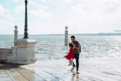 A father joyfully swings his daughter by the sea on a bright day, capturing a playful moment.